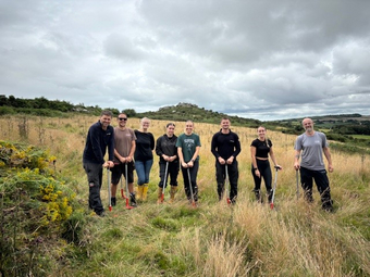 Eight people stand in a coastal field and lean on shovels.