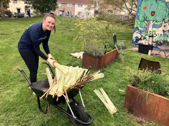 A woman smiles at the camera next to a wheelbarrow of gardening waste.